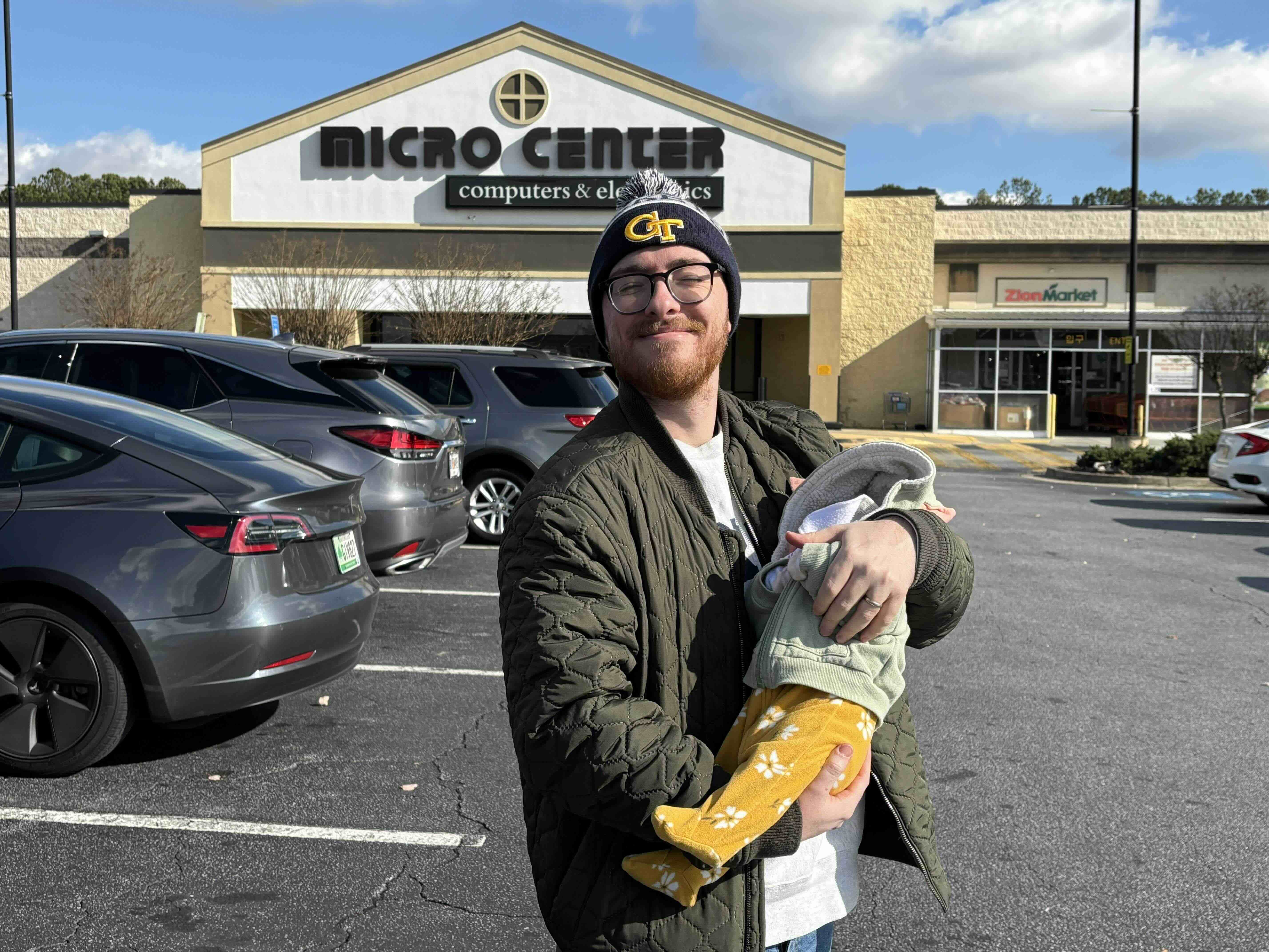 Me, holding my infant child and smiling in the parking lot of a Microcenter. I am wearing a coat and winter hat. My kid is wearing a Grogu jacket from The Mandolorian and a yellow onesie with flowers.